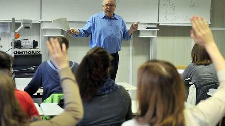Ein Lehrer steht gestikulierend und lachend vor einer Schulklasse, die Schüler melden sich.