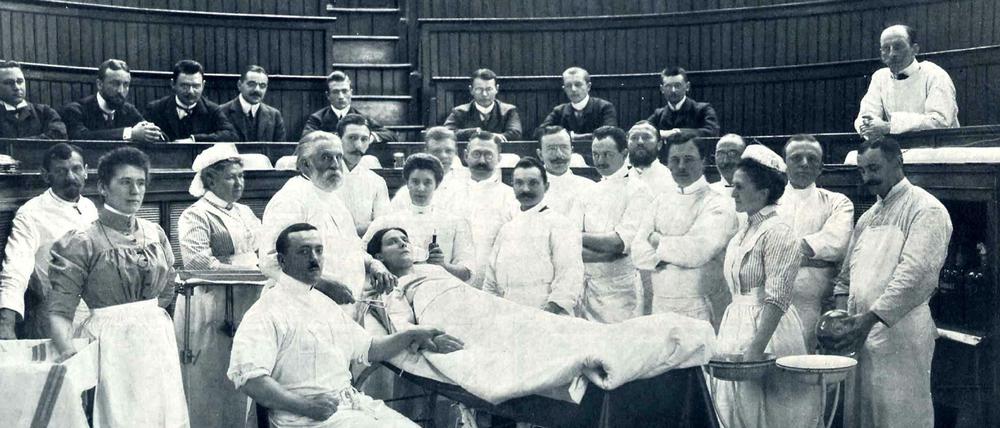 Der Chirurg Ernst von Bergmann mit grauem Bart steht im Operationssaal am Kopfende einer liegenden Patientin. Der Arzt ist umgeben von Assistenzärzten, Pflegern und Krankenschwestern.