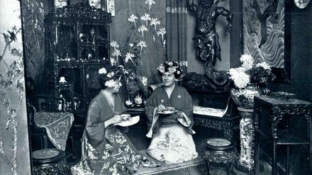 Zwei Damen sitzen bekleidet in Kimonos in einem japanisch eingerichteten Zimmer und halten Teetassen in der Hand.