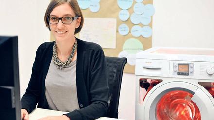Eine junge Frau sitzt am Computer, im Hintergrund steht eine Waschmaschine.