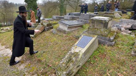 Ein Rabbiner zeigt auf einem jüdischen Friedhof auf einen umgekippten Grabstein.