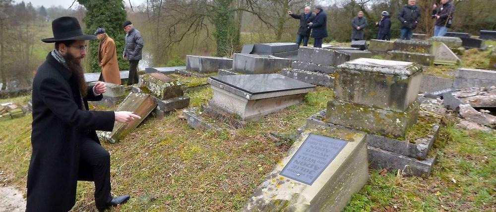 Ein Rabbiner zeigt auf einem jüdischen Friedhof auf einen umgekippten Grabstein.