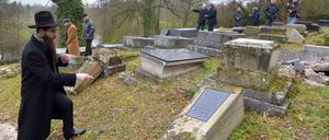Ein Rabbiner zeigt auf einem jüdischen Friedhof auf einen umgekippten Grabstein.