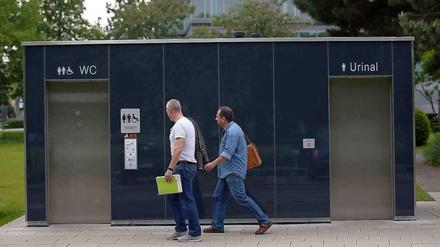 Zwei Männer gehen in Köln an einer öffentlichen Toilette vorbei. Frauen müssen für die Benutzung dieser Toilettenanlagen 50 Cent zahlen, während Männer gratis das Urinal benutzen dürfen.