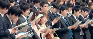 Studenten stehen in einem Saal und blicken lächelnd auf Dokumente, die sie in Händen halten.