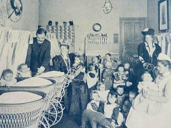 Ein Mann und drei Frauen in altmodischer Kleidung kümmern sich in einem Zimmer um Kleinkinder, vor einer Gruppe von Kindern steht ein Stoffelefant auf Rädern.