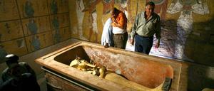 Drei Männer stehen in einem mit Wandbildern geschmückten Raum neben einem Sarkophag.