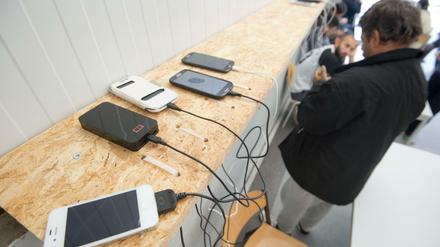 Auf einem Holzregal liegen Mobiltelefone, die aufgeladen werden, im Hintergrund sind wartende Männer zu sehen.