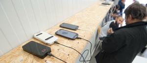 Auf einem Holzregal liegen Mobiltelefone, die aufgeladen werden, im Hintergrund sind wartende Männer zu sehen.