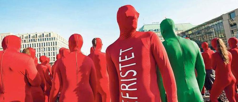 Menschen in roten Ganzkörperanzügen mit der Aufschrift Befristet stehen auf einem Platz.
