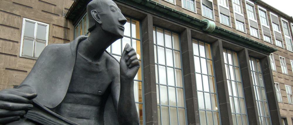 Eine Skulptur vor einem Universitätsgebäude stellt einen sinnierenden Mann mit einem aufgeschlagenen Buch auf den Knien dar.