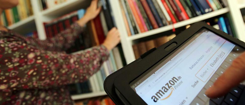 Auf einem Tablet-Computer sucht ein Mann bei dem Online-Versandhaus Amazon nach Literatur, während seine Frau ein Buch aus dem Bücherregal nimmt. 