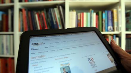 Auf einem Tablet-Computer ist die Seite des Online-Versandhaus Amazon vor einem Bücherregal geöffnet.