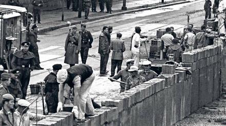 Bauarbeiter bauen eine Mauer, sie werden von bewaffneten Uniformierten bewacht.