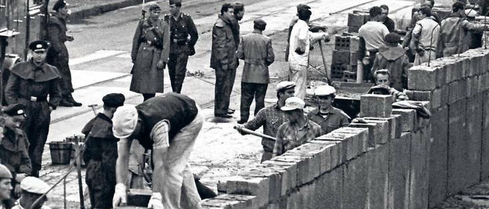 Bauarbeiter bauen eine Mauer, sie werden von bewaffneten Uniformierten bewacht.