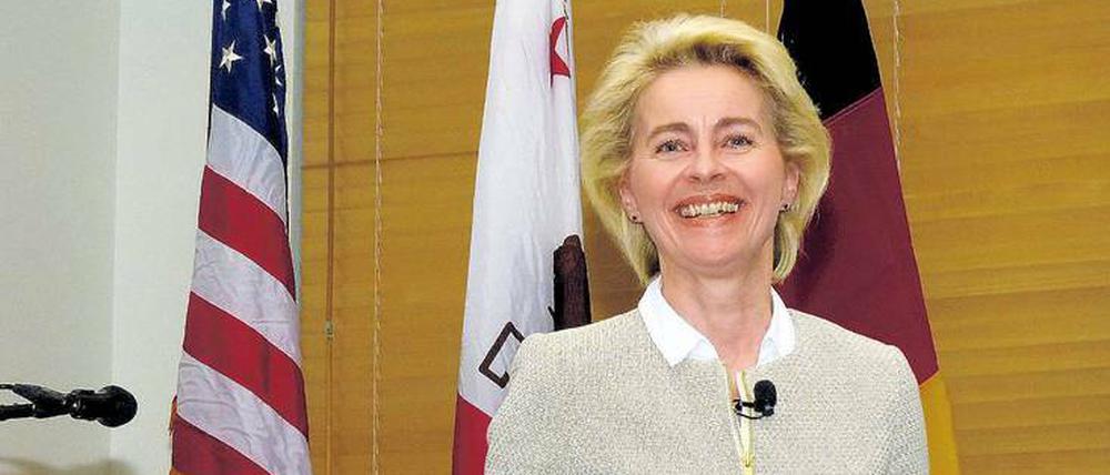 Eine Frau steht mit einem strahlenden Lächeln im Gesicht vor einer Wand und gestikuliert mit ihren Händen.