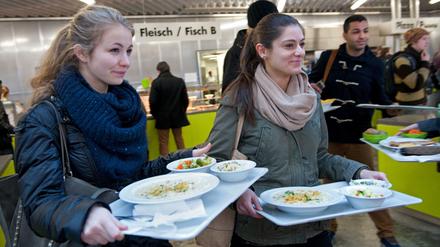 Zwei junge Frauen gehen mit beladenen Mensa-Tabletts durch einen Raum.