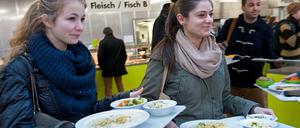 Zwei junge Frauen gehen mit beladenen Mensa-Tabletts durch einen Raum.