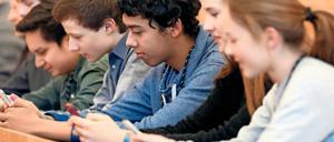 Jugendliche sitzen in einer Reihe an einem Arbeitstisch und hantieren mit ihren Smartphones.