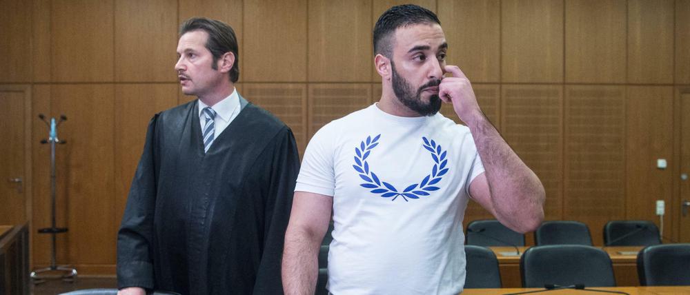 Mit einem weißen T-Shirt bekleidet steht der Angeklagte Aria L. in einen Hochsicherheitssaal des Oberlandesgerichts in Frankfurt am Main neben seinem Anwalt Andreas Bensch.