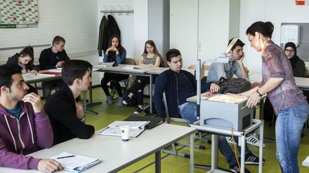 Junge Frauen und Männer sitzen im Klassenraum, vor ihnen steht eine Lehrerin.