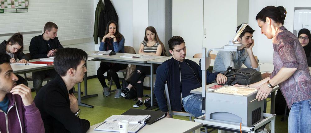 Junge Frauen und Männer sitzen im Klassenraum, vor ihnen steht eine Lehrerin.