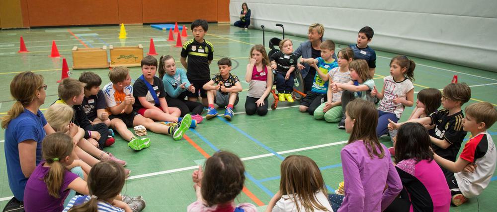 Lehrkräfte und Schulkinder in Sportbekleidung sitzen in einer Turnhalle in einem Kreis und sprechen miteinander. Ein Kind sitzt im Rollstuhl.