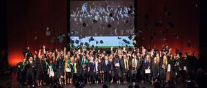 Studierende stehen in einem Saal und werfen schwarze Hüte in die Luft.