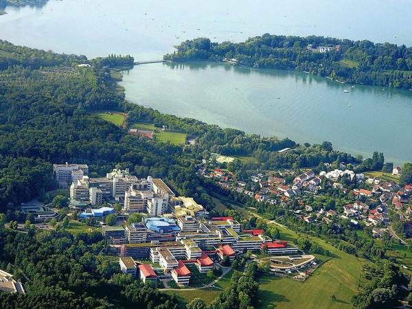 Ein Universitäts-Campus mit moderner Hochhausarchitektur liegt in der Nähe eines Seeufers und inmitten von Wiesen und Wäldern.