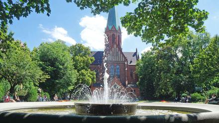 Blick von Westen auf die Grünanlage des Ludwigkirchplatzes in Berlin-Wilmersdorf mit dem Brunnen vorn und der katholischen Kirche St. Ludwig im Hintergrund.