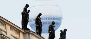 Auf dem Dachsims eines Gebäudes stehen vier Skulpturen, im Hintergrund die Aussichtskugel eines Fernsehturms.