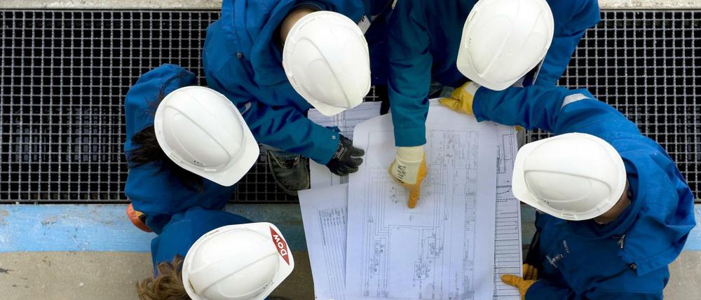 Eine Gruppe von Ingenieuren mit Bauhelmen auf dem Kopf beugt sich über Baupläne.