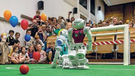 Roboter spielen Fußball, Kinder und Erwachsene schauen ihnen dabei zu.