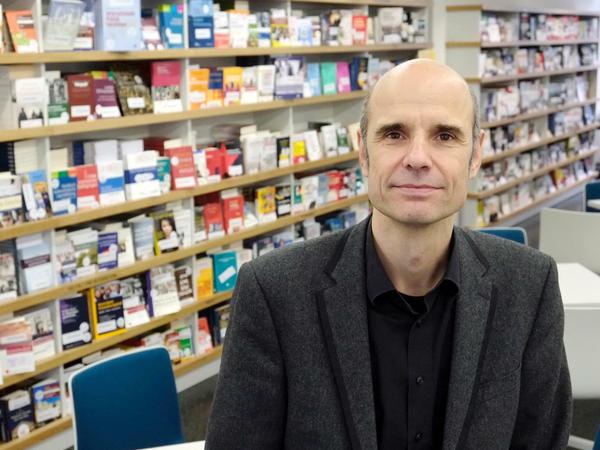 Ein Mann steht vor einem Bücherregal und schaut in die Kamera.