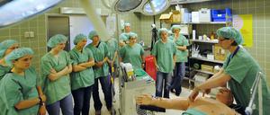 Studierenden in grünen Kitteln stehen in einem Operationssaal um eine Übungspuppe herum.