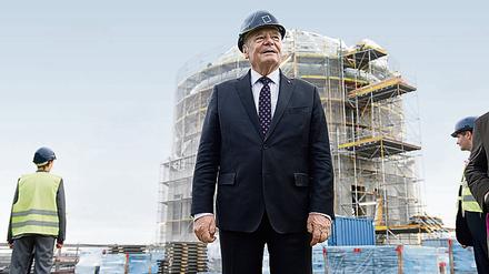 Bundespräsident Joachim Gauck steht mit herkömmlichen Arbeitsschuhen und Helm bekleidet auf dem Dach des Gebäudes vor der Kuppel. 