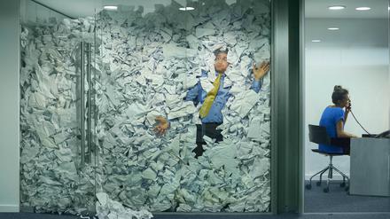 A businessman is trapped in his glass office by a surplus of discarded ideas on paper . His colleague in the next office is working more efficiently and is oblivious to him being trapped .