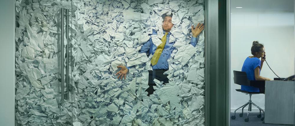 A businessman is trapped in his glass office by a surplus of discarded ideas on paper . His colleague in the next office is working more efficiently and is oblivious to him being trapped .