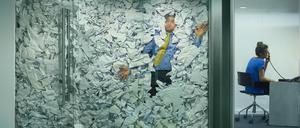 A businessman is trapped in his glass office by a surplus of discarded ideas on paper . His colleague in the next office is working more efficiently and is oblivious to him being trapped .
