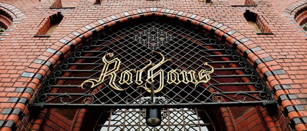 Portal und rote Backsteinfassade des Rathauses Lichtenberg mit historischem "Rathaus"-Schild am Gitter.