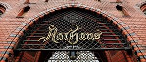 Portal und rote Backsteinfassade des Rathauses Lichtenberg mit historischem "Rathaus"-Schild am Gitter.