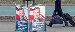 Um einen Baum herum stehen Wahlplakate, ein Mann, der einen Rollkoffer hinter sich herzieht, geht daran vorbei.