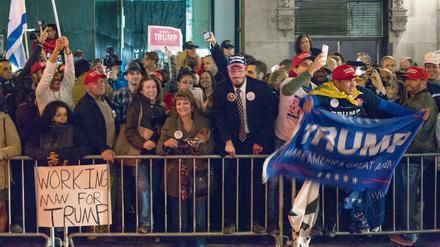 Menschen, die Transparente mit der Aufschrift Trump hochhalten, stehen jubelnd hinter einer Absperrung. Im Vordergrund ist ein Schild mit der Aufschrift "Working Man For Trump" zu sehen.