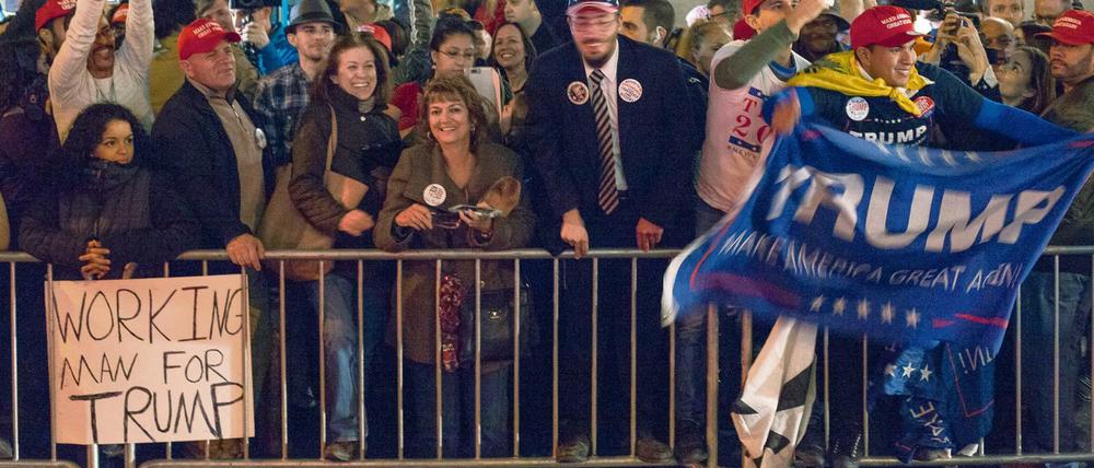 Menschen, die Transparente mit der Aufschrift Trump hochhalten, stehen jubelnd hinter einer Absperrung. Im Vordergrund ist ein Schild mit der Aufschrift "Working Man For Trump" zu sehen.
