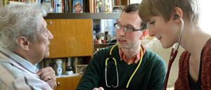Ein Arzt und eine Medizinstudentin im Gespräch mit einer älteren Patientin in deren Wohnzimmer.