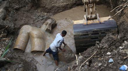 Das Fragment einer Statue liegt im Erdreich und in einem Wasserloch, ein Arbeiter versucht neben einer Baggerschaufel ein weiteres Fragment mit den Händen zu bewegen.