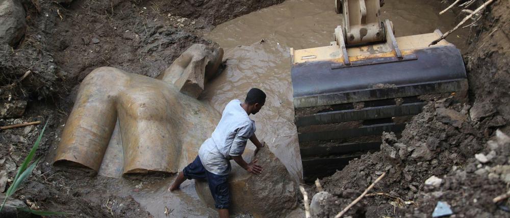 Das Fragment einer Statue liegt im Erdreich und in einem Wasserloch, ein Arbeiter versucht neben einer Baggerschaufel ein weiteres Fragment mit den Händen zu bewegen.