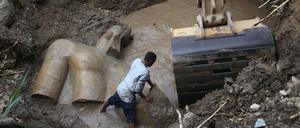 Das Fragment einer Statue liegt im Erdreich und in einem Wasserloch, ein Arbeiter versucht neben einer Baggerschaufel ein weiteres Fragment mit den Händen zu bewegen.