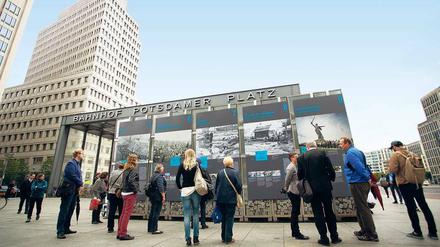 Auf einem von Hochhäusern gesäumten Platz und dem Eingang des Bahnhofs Potsdamer Platz steht eine Freiluft-Ausstellung, davor stehen Menschen, die die Tafeln der Ausstellung lesen.