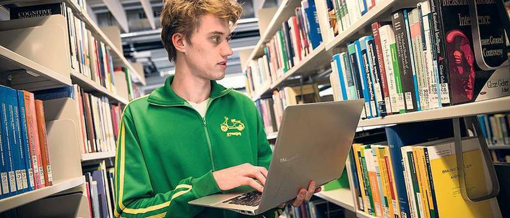 Ein Student steht mit aufgeklapptem Laptop zwischen den Regalen einer Bibliothek.